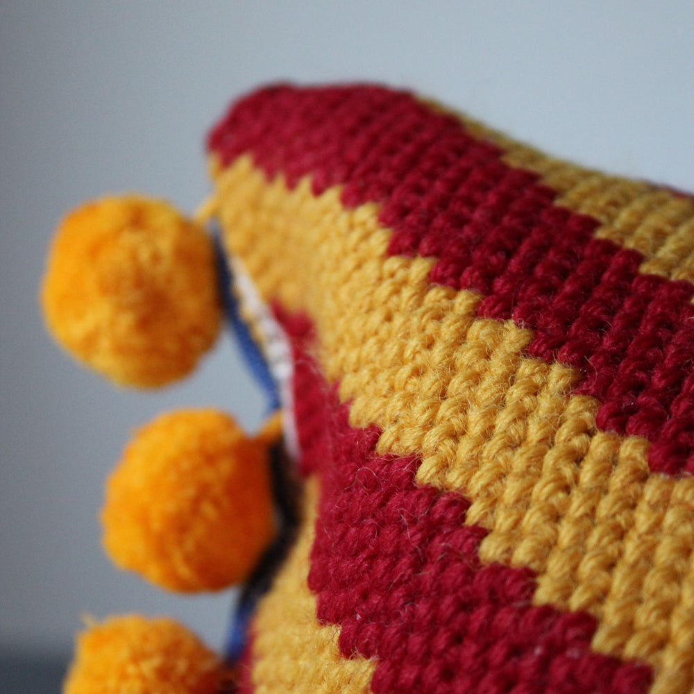 Close-up of a cross stitch tapestry tuck shop caramel jacqui pearce copyright  item with red and yellow pattern and orange pom-poms on a blurred background