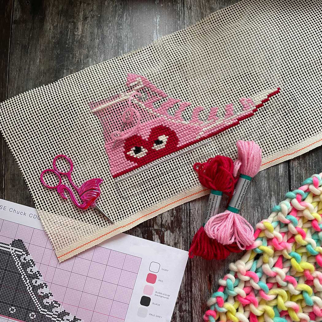 Cross-stitching project with pink heart shoe design on fabric, surrounded by yarn and patterns.