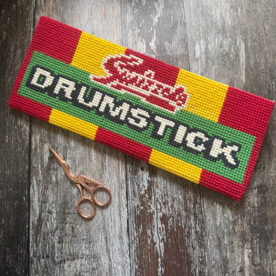 Cross-stitched patch with 'Swizzles Drumstick' design on a wooden surface with scissors.