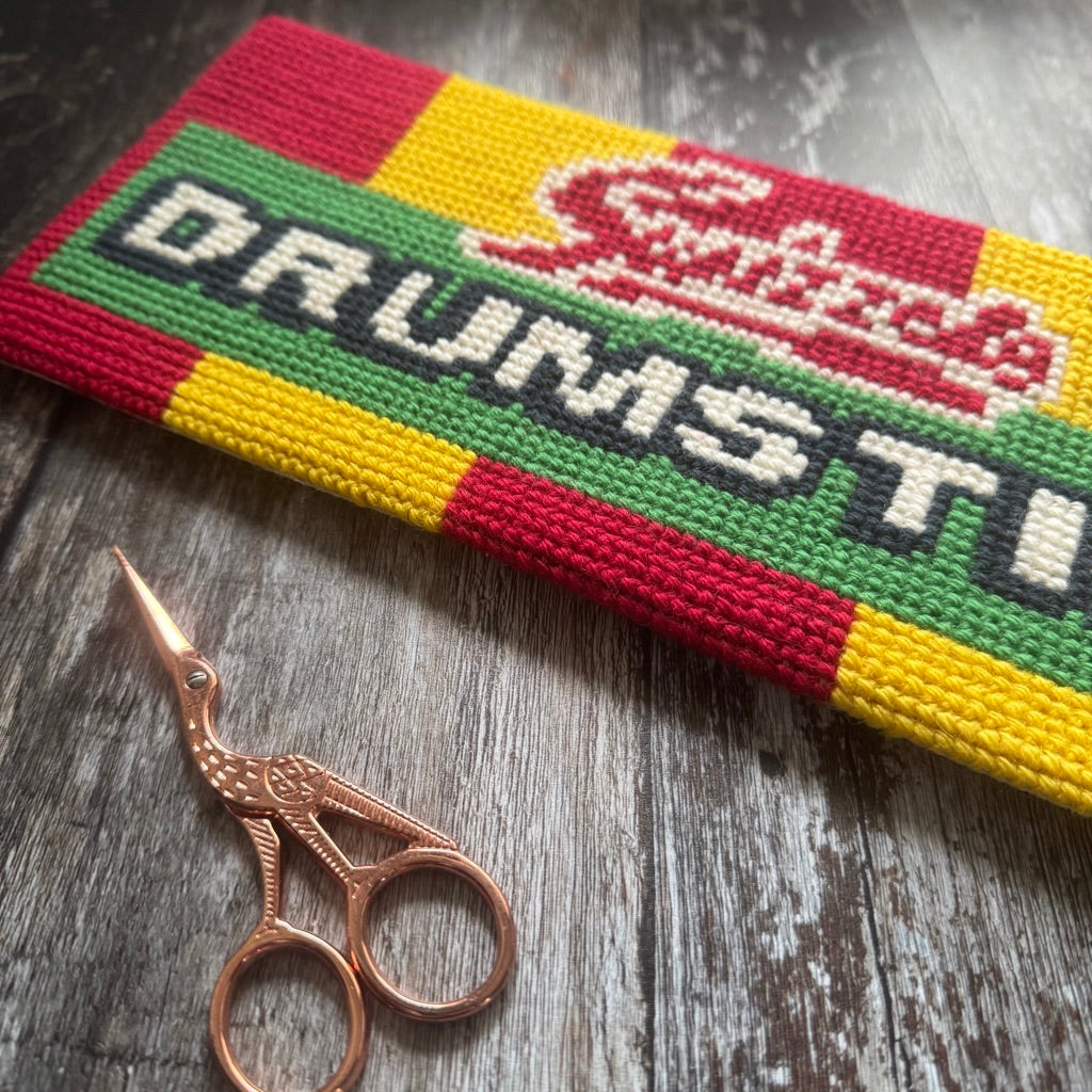 Cross-stitched drumstick lolly with colorful design and text, accompanied by a pair of decorative scissors on a wooden surface.
