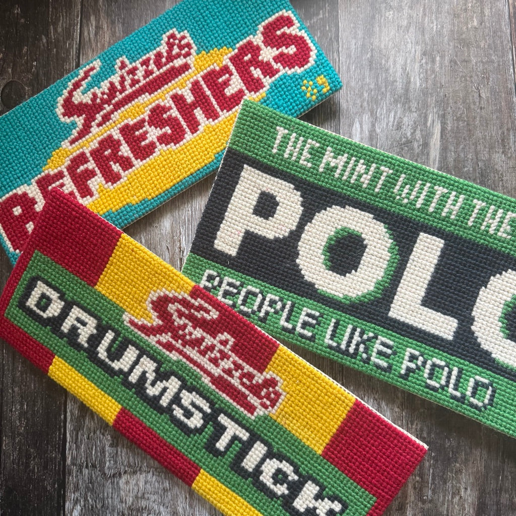 Three colorful tuck shop cross stitch designs with text on a wooden surface